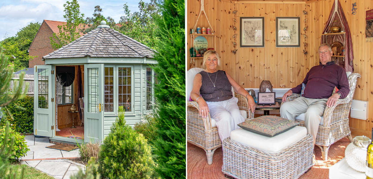 A close up view of the summerhouse on the right surrounded by green bushes and plant foliage. On the right is Chris & Moira sitting on the comfy chairs looking directly at the camera smiling.