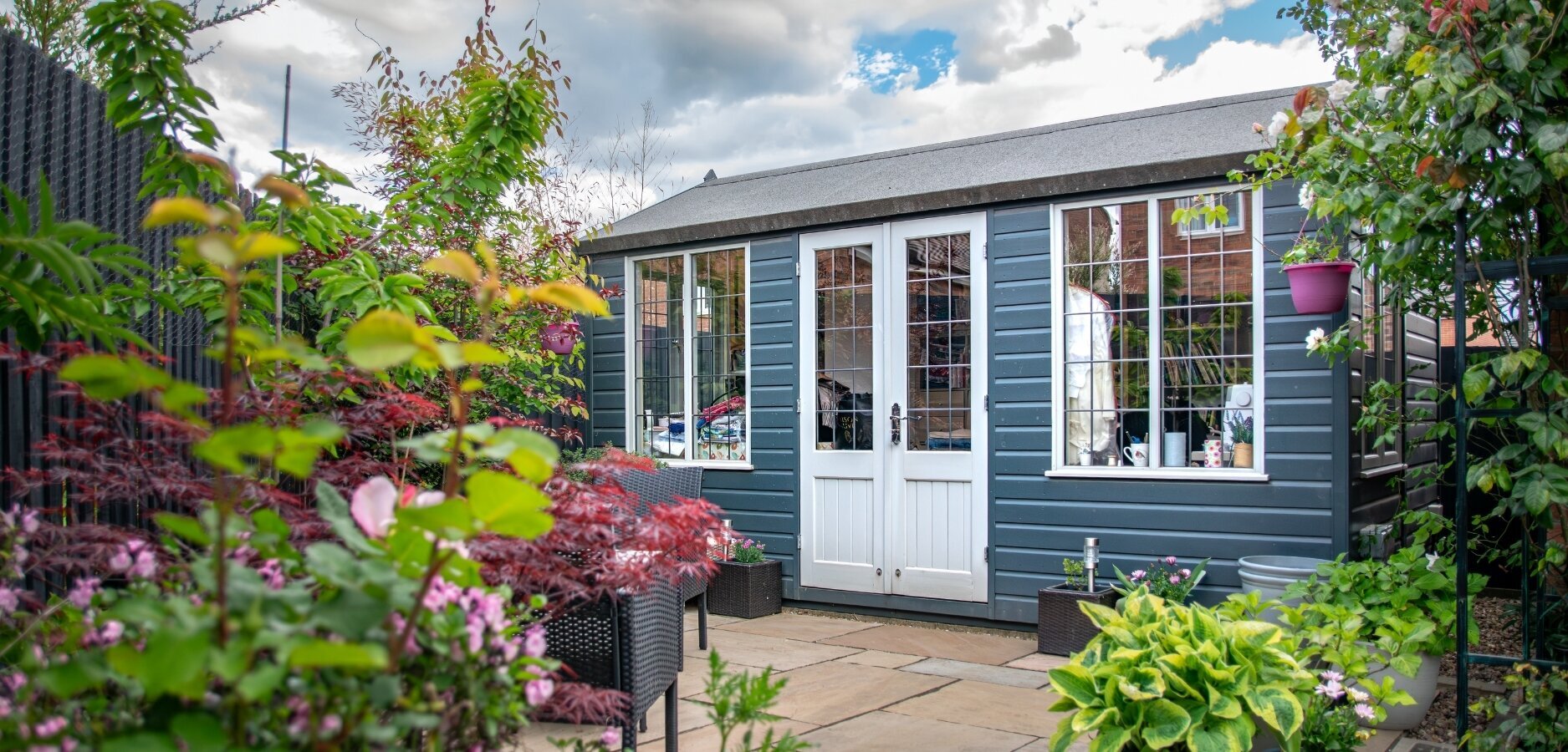 The quaint traditional style summerhouse painted in dark grey with light contrasting windows. Surrounded by vibrant tree foliage.