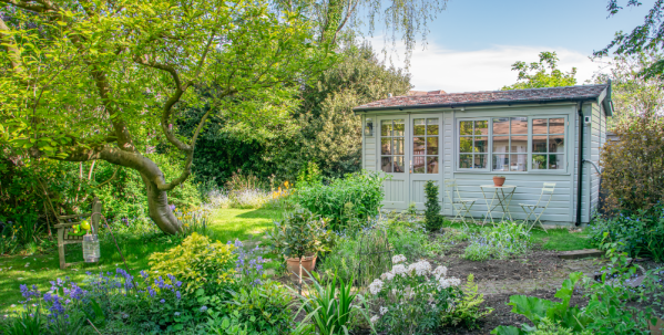 Langham Studio in a mature garden A sage green timber garden room surrounded by lush planting, mature trees and colourful flower beds, with a small bistro table outside.