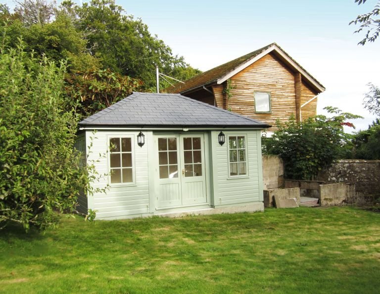 A small garden room with a grey-slate-composite tiled roof and Georgian windows. The building has inset doors and corner boards and black guttering along the fascia boards. 