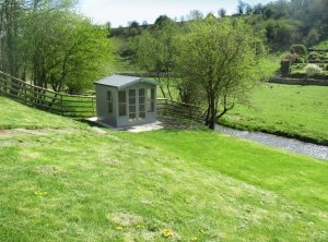 2.4 x 2.4m Blakeney Summerhouse painted in Exterior Lizard with Shiplap Cladding and an Overhanging Apex Roof pictured in an idyllic spot next to a river