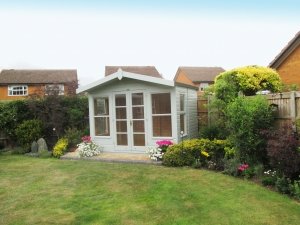A traditional summerhouse in sage paint with an apex roof and chalet-style overhang. Large windows have been placed either side double doors.