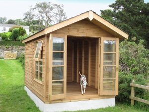 A traditional blakeney summerhouse with windows and doors open. A dog is leaving the building and heading towards the camera. The building has an apex roof and is painted in a natural light oak preservative which allows the natural wood to show through. 