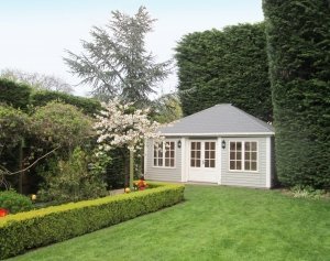 A large garden building pictured in an attractive garden with tall conifer hedges and decorative shrubbery. The building has a hipped roof covered in slate composite tiles and is clad with rustic weatherboard cladding. It is painted in both a light grey colour called Pebble and ivory on the doors and window frames. 