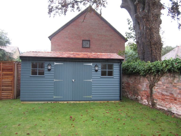 A large superior shed in a dark slate paint and shiplap cladding. It has an apex roof covered with cedar shingles, double door entry and georgian windows. 