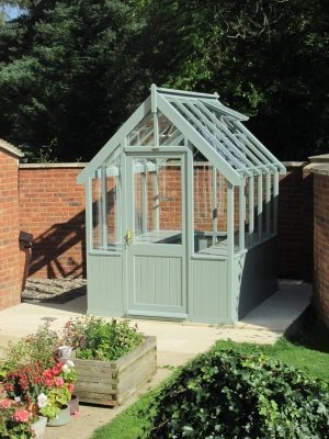 A small greenhouse painted in the exterior shade of lizard on a garden patio within a walled garden. 