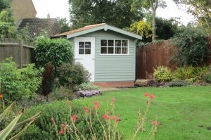 An attractive shed situated in the corner of a decorative garden with red flowers and shrubbery. The shed has an apex roof covered with cedar shingles and a slight overhang. The door and windows are painted ivory while the rest is painted in Lizard. The shed is clad with rustic weatherboard. 