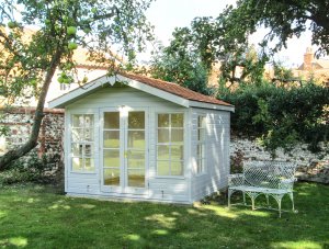 A chalet-style blakeney summerhouse in a stunning walled garden with trees and an antique outdoor bench. The summerhouse has an apex roof covered with cedar shingles and smooth shiplap cladding painted in ivory. 
