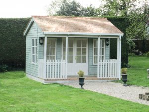 A small garden room with a veranda beneath the overhang of the apex roof covered in cedar shingles. The exterior is clad with rustic weatherboard cladding painted Lizard exterior paint with guttering added to the fascia boards. 