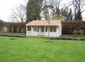 A large and attractive pavilion garden room situated in an attractive garden with a veranda beneath an overhang on the apex roof.