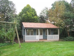 A large garden room pictured in a wooded garden with a children's swing beside it. The building has a large veranda beneath the overhang of the apex roof which is covered in cedar shingles. Guttering has also been added to the fascia boards. 