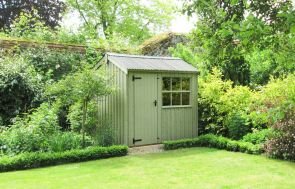 An image showing the Felbrigg National Trust shed pictured in a leafy garden with an abundance of shrubbery. The shed has an apex roof covered with corrugated material and vertically-sawn, rustic-cut cladding