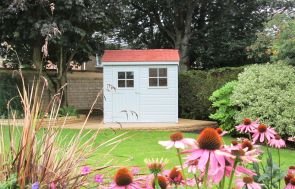 A small superior shed in a large garden with pink flowers in the foreground. The shed has an apex roof covered with terracotta tiles and smooth shiplap cladding painted in sage exterior paint. 