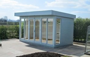 An image of the salthouse studio on a patio area overlooking a rural landscape. The building has a pent roof and smooth shiplap cladding painted in the exterior shade of verdigris. The building has six fully-glazed windows and fully-glazed double doors, meaning that the front of the building is mainly glass. The visible interior has light painted walls and a natural wood floor. 
