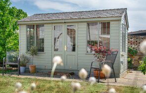 Traditional style apex summerhouse painted in a soft green post installation surrounded by greenery and some potted plants.
