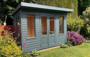 A Thornham summerhouse which is a traditional style garden building with windows on each side and a pent roof. In a bright green garden with some flowers