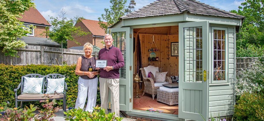 Chris and Moira standing outside their beautiful hexagonal summerhouse with the doors open to show the cosy interior dressing surrounded by the couples immaculate green foliage, small trees and flowering plants.