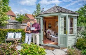 Chris and Moira standing outside their beautiful hexagonal summerhouse with the doors open to show the cosy interior dressing surrounded by the couples immaculate green foliage, small trees and flowering plants.