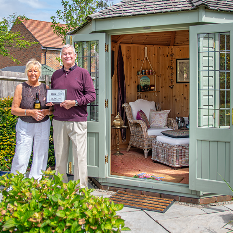 Chris and Moira standing outside their beautiful hexagonal summerhouse with the doors open to show the cosy interior dressing surrounded by the couples immaculate green foliage, small trees and flowering plants.