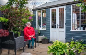 2.4 x 4.2m Holkham Summerhouse painted in Slate with Apex Roof the customer is sitting outside the front of the building on her garden furniture smiling. There is lots of green trees and foliage in the background.