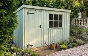 Pent roof traditional shed with rustic vertical sawn cladding, georgian windows, and a black corrugated roof with a flower bed around the outside with flowering plants