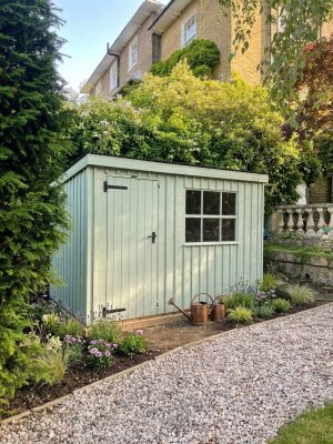Pent roof traditional shed with rustic vertical sawn cladding, georgian windows, and a black corrugated roof with a flower bed around the outside with flowering plants